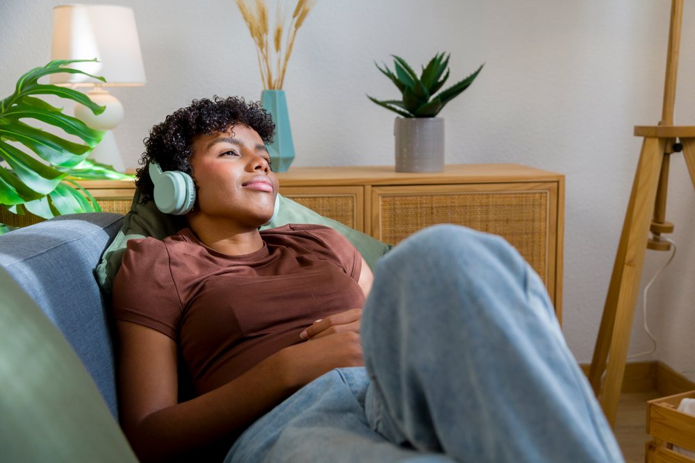Woman relaxing on sofa wearing headphones in cozy modern home interior with plants and wooden furniture.