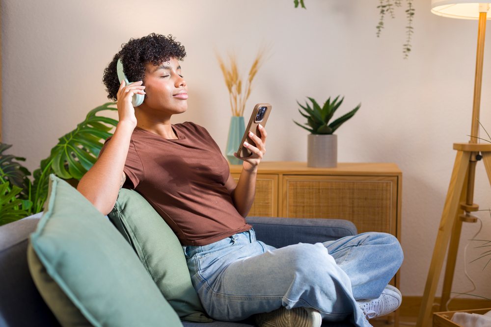 Young woman with curly hair seated on a couch at home listens to music with headphones and holds a smartphone.