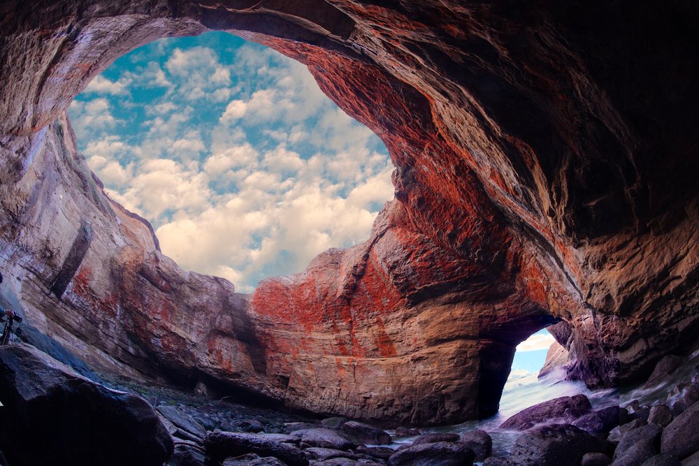 Devils Punch bowl cave with rocky formations and ocean water visible through a large opening under a cloudy sky, Oregon, USA.