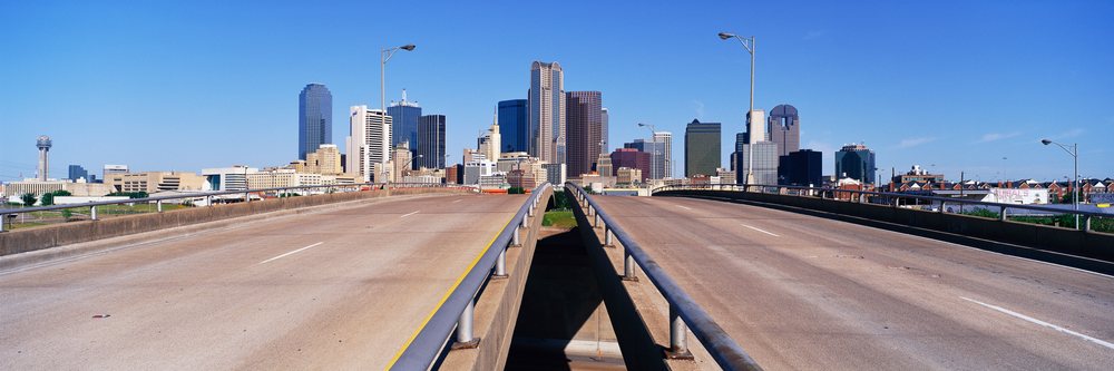 Empty highway bridge foreground with the distinctive Downtown Dallas skyline under a clear blue sky.