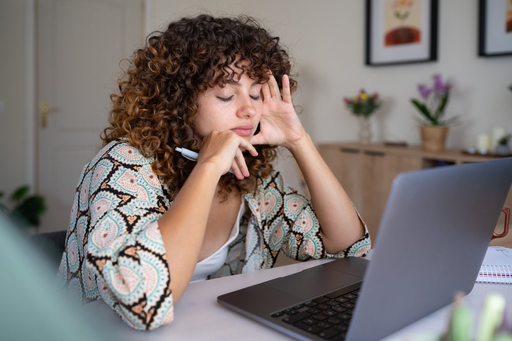 Woman with curly hair sitting at a desk using a laptop and holding a pen in a home interior, Spain.