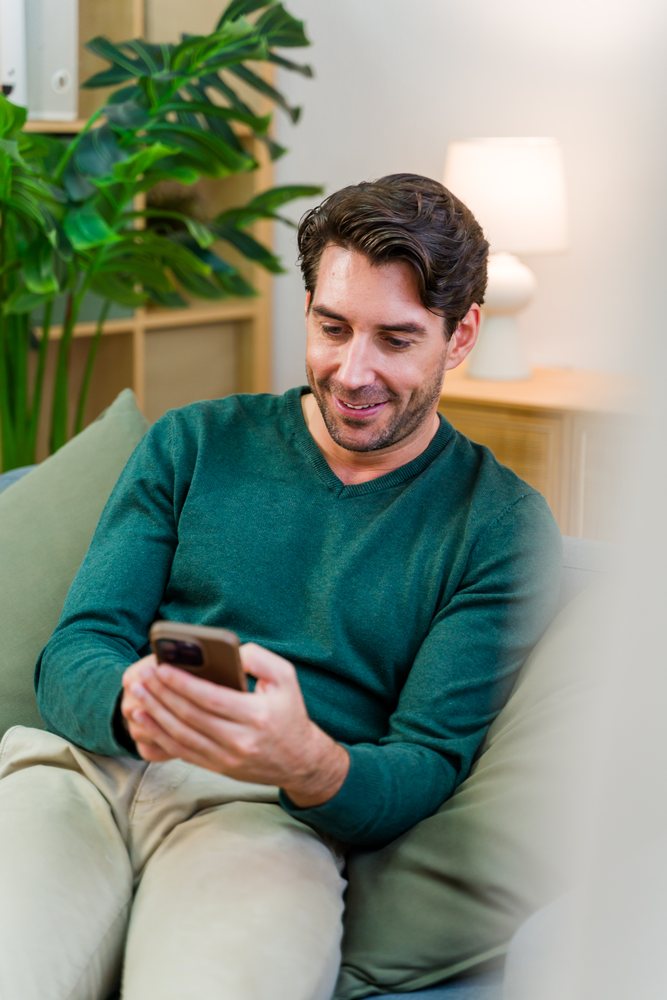 Man wearing green sweater and beige pants sitting on sofa smiling while holding smartphone in modern living room with indoor plant and table lamp, Spain.