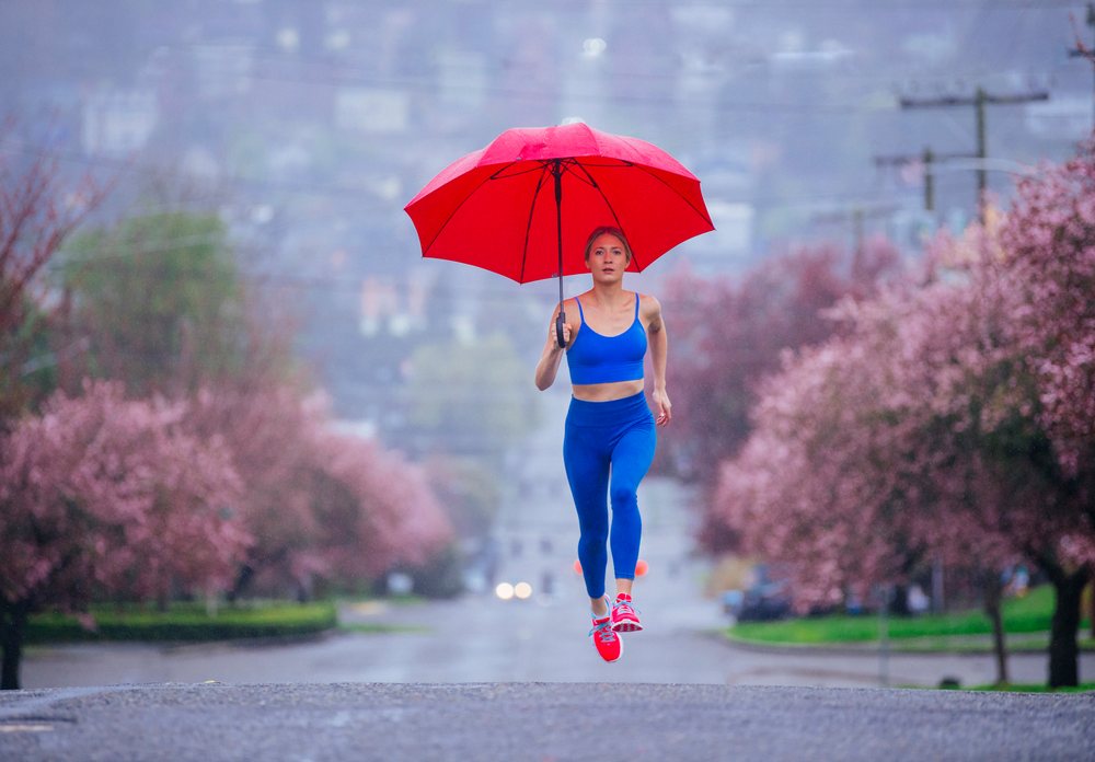 Woman running on an urban street in spring rain holding a red umbrella beside pink flowering trees, USA.