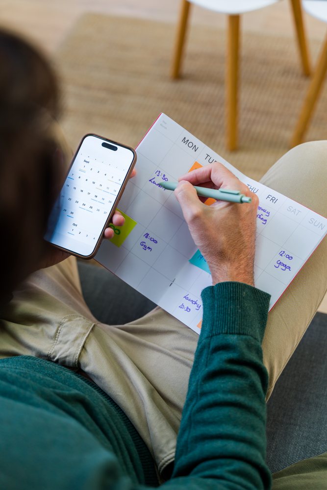 Person indoors writing appointments in a weekly planner while referencing a mobile phone calendar, Spain.