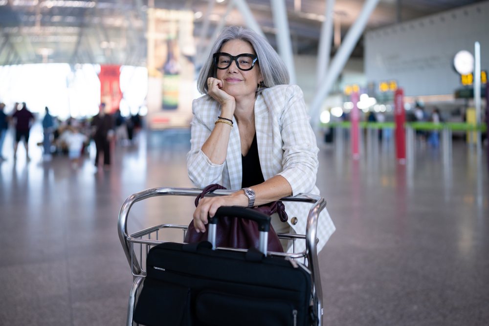 Businesswoman traveler with gray hair leans on luggage trolley in a spacious airport terminal, Spain, Madrid.