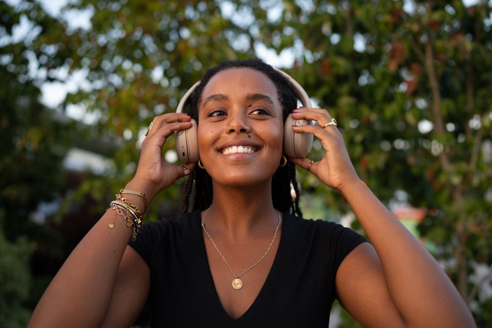 A smiling woman adjusts her wireless audio device outdoors amid greenery, urban, Santiago De Compostela, Spain.