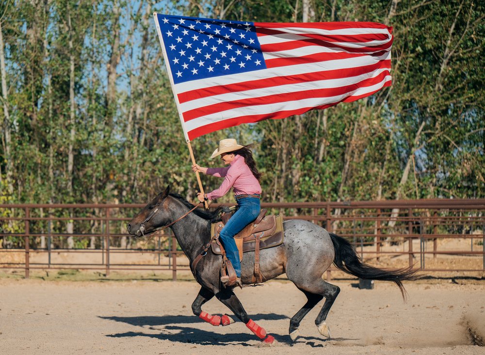 Woman wearing cowboy hat gallops a horse in an outdoor arena while holding a large American flag on a pole, Southwest USA.