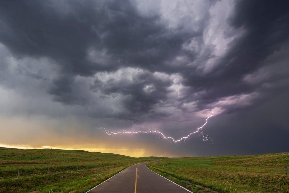 Lightning strikes beneath dark storm clouds over a two-lane road flanked by grassland and fence posts in the Nebraska Sandhills during a storm chase at twilight, Sutherland, USA.
