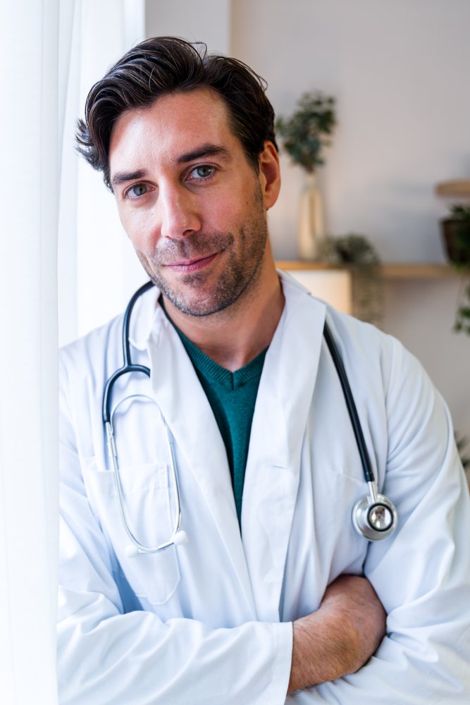Male doctor wearing a white coat and stethoscope stands with arms crossed inside a medical examination room indoors, Spain.