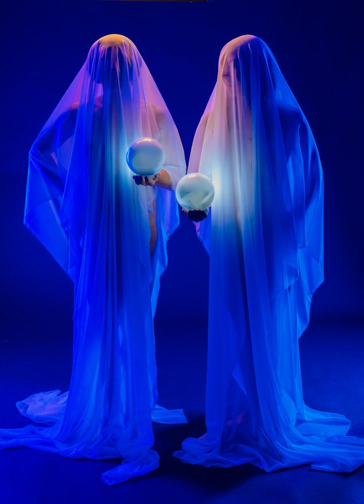 Two people draped in white translucent fabric hold glowing spheres in a blue-lit studio, ghosts, USA.