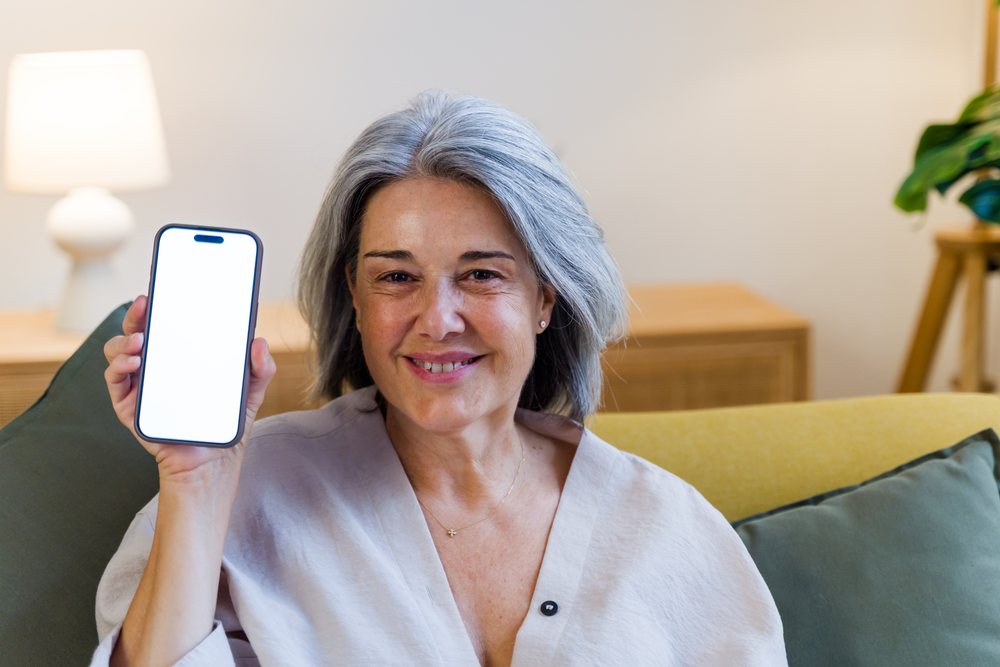 Mature woman with gray hair smiles while holding a smartphone with a blank screen in a cozy living room, Spain.