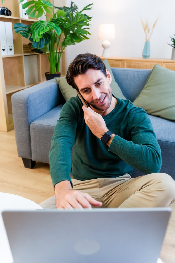 Man in green sweater sitting on the floor talking on the phone and using a laptop in a living room, Spain.