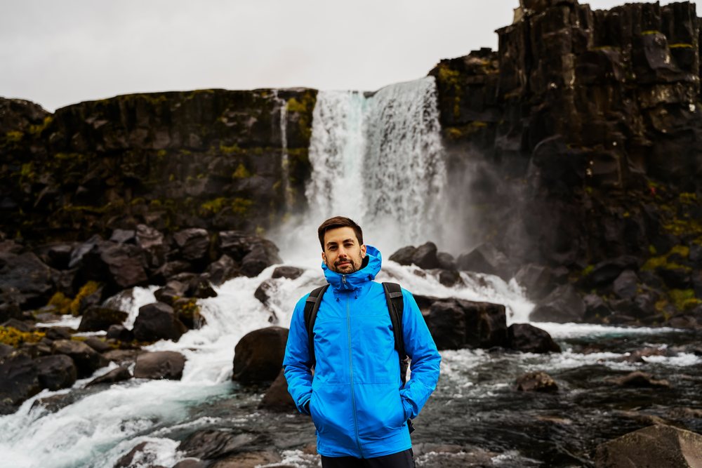 A man wearing a blue jacket and backpack stands on rocky terrain with a large waterfall flowing behind him, Iceland.
