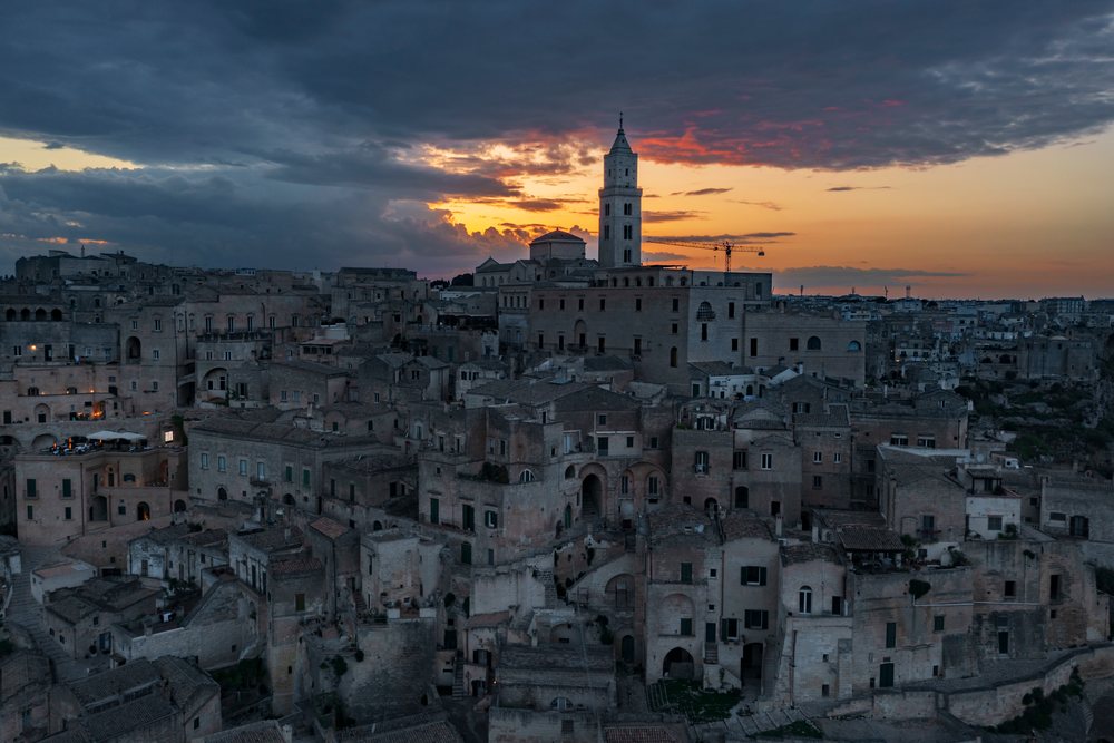 Sunset illuminates Sasso Caveoso, highlighting historic stone buildings and a prominent tower under a cloudy sky, Matera, Basilicata, Italy.