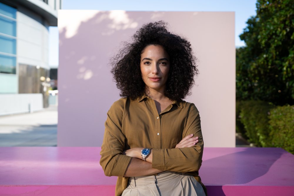 Woman with curly hair and crossed arms stands confidently outdoors against a pink wall in natural sunlight, Madrid, Spain.