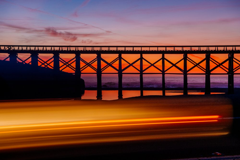 Silhouette of a wooden bridge spanning the ocean coast at sunset with vehicle light trails on the road below in northern Oregon and, California, USA.