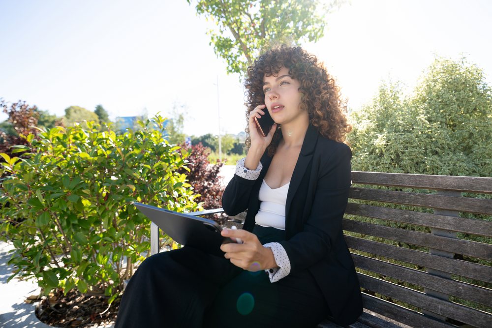 Woman with curly hair wearing a business outfit talks on phone while using a tablet on a bench in an outdoor park during daytime, Spain.