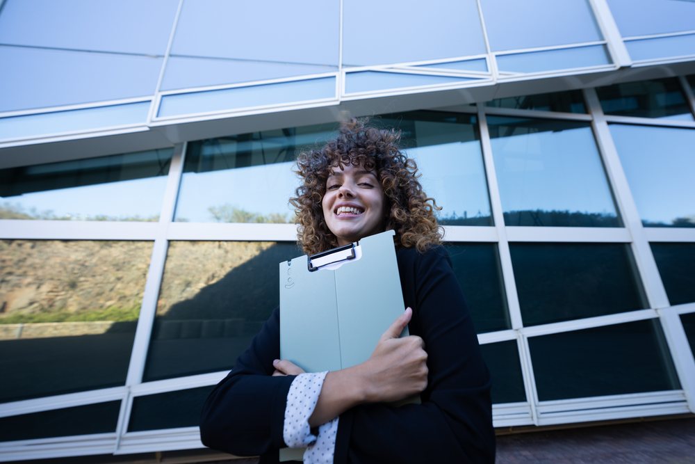 Businesswoman holding a folder and smiling in front of a modern glass building, business trip, Spain, Santiago De Compostela.