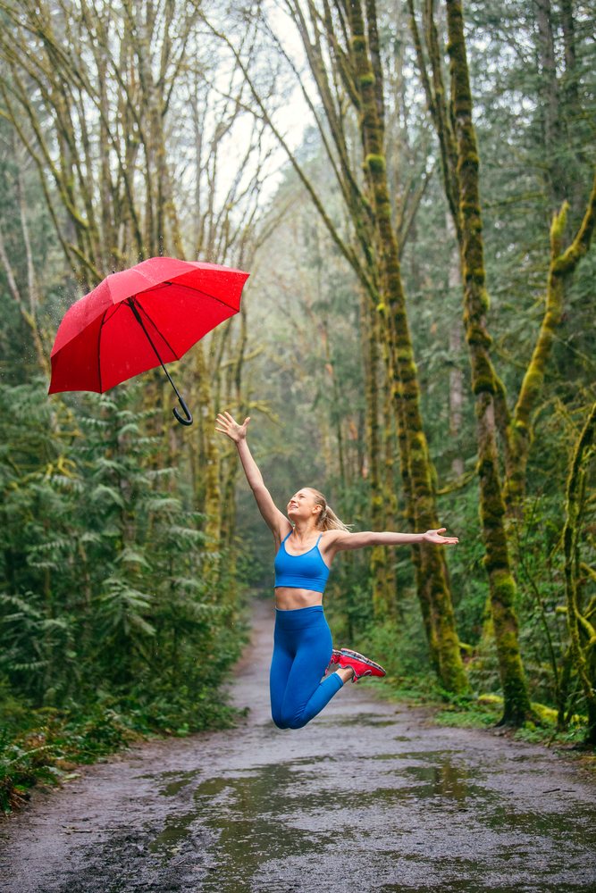 Woman joyfully jumping and tossing a red umbrella on a rainy forest trail with moss-covered trees and wet ground, USA.