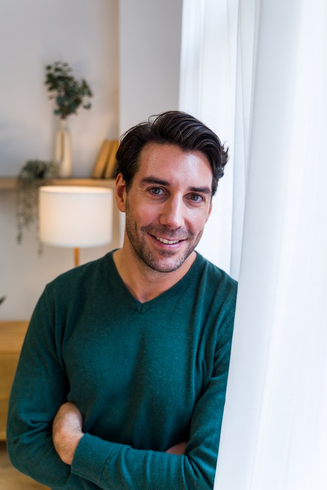 Man smiling indoors with crossed arms by a white curtain near a lamp and indoor plants on a wooden shelf with books, Spain.