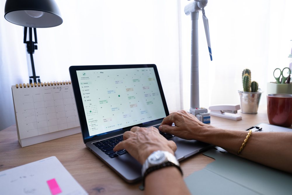 Hands typing on a laptop with a digital calendar and paper calendar on a desk in a bright home office workspace, Spain.