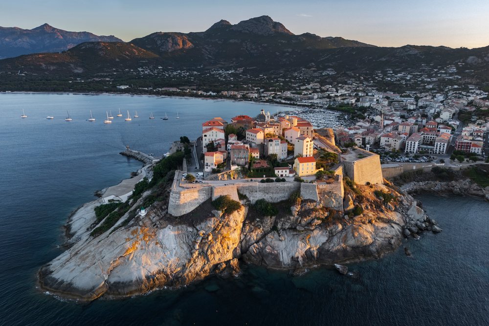 Stone citadel on a rocky promontory with red-roofed buildings, boats in harbor, mountains, and sea at Sunset, Travel Photography, Calvi, Corsica, France.