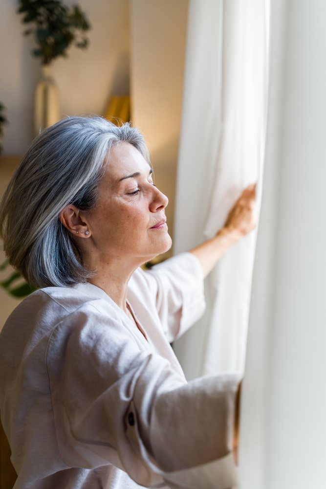 Senior woman with gray hair opens a curtain to let natural sunlight indoors at her home, Spain.