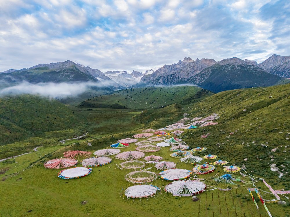 Multiple circular Tibetan Buddhist prayer flags are laid out on a grassy valley floor with rocky mountains and partly cloudy skies in Kham Tibet, China, Zhuqing, Sichuan Province, People's Republic of China.