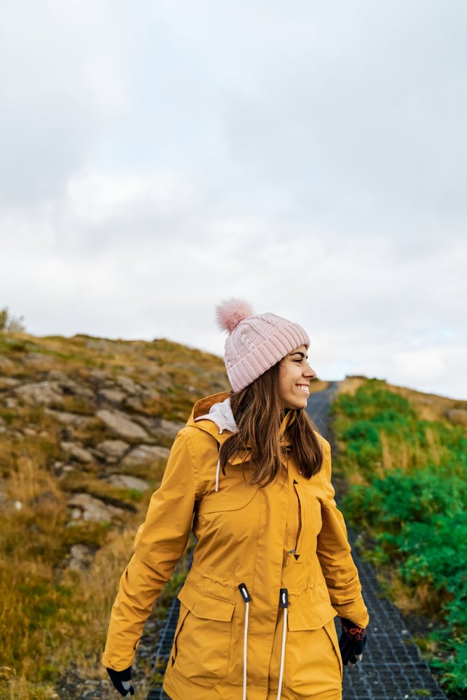 A woman in a yellow jacket and pink knit hat smiles while walking along a rocky path in nature, Iceland.