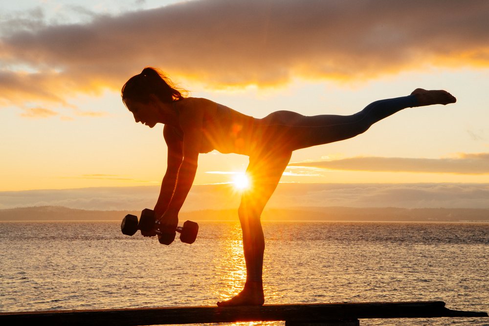 Woman holding dumbbells balances on one leg at sunset over water, WA, USA.