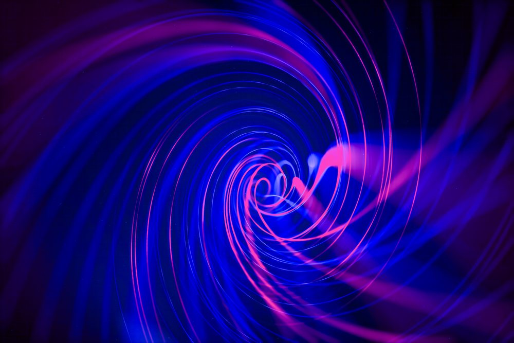An abstract swirling vortex of blue and pink light trails creating a dynamic glowing pattern on a dark background.