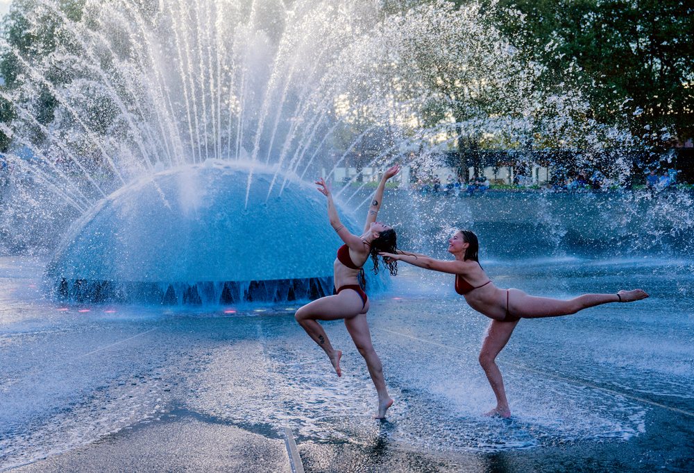 Two women in swimwear performing an acrobatic dance with water splashing from a fountain in an outdoor setting, partner, acrobatics, yoga, USA.