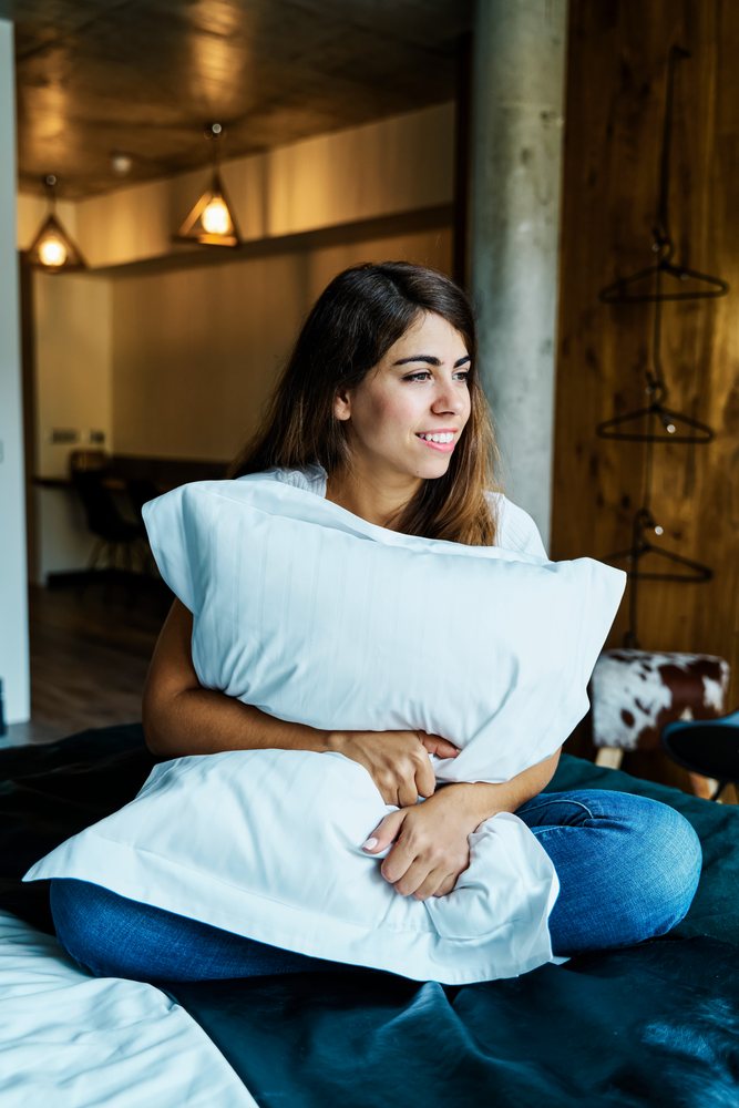 A woman sitting on a bed hugging a pillow in a bedroom interior