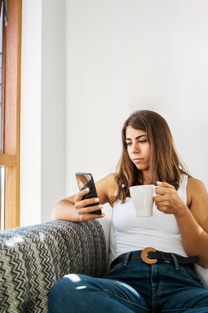 A white woman relaxes on a couch using a smartphone and holding