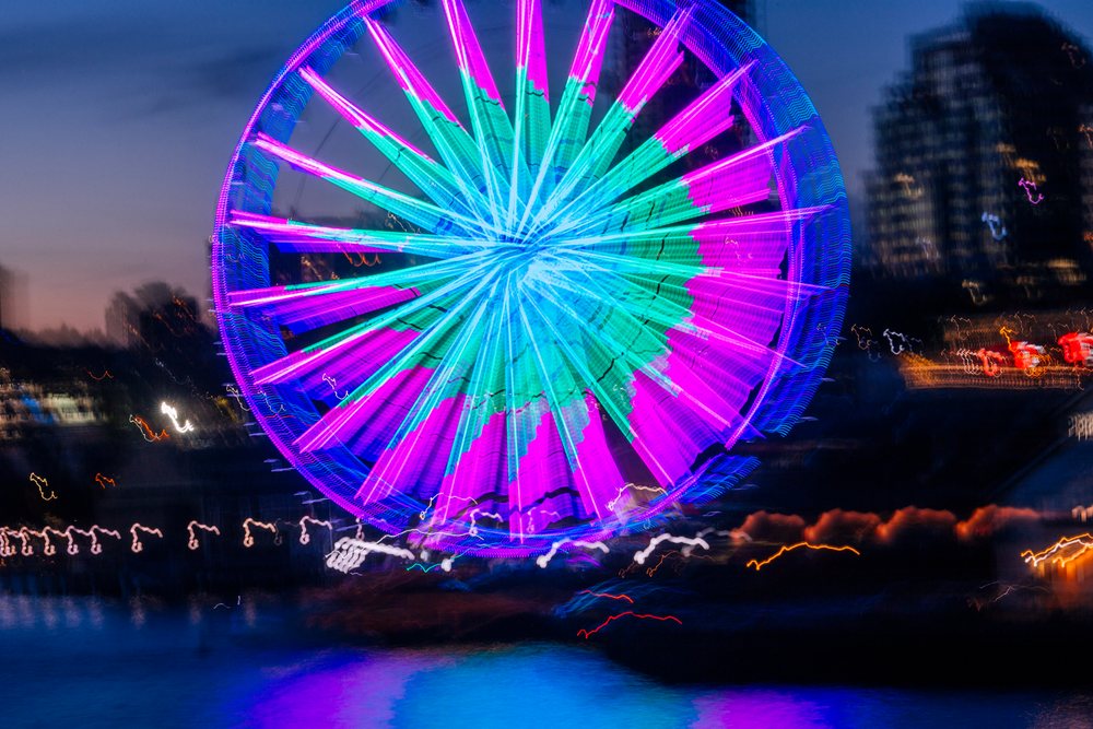 Neon-lit ferris wheel at night reflecting over water in Seattle cityscape with light trails and urban skyline, WA, USA.