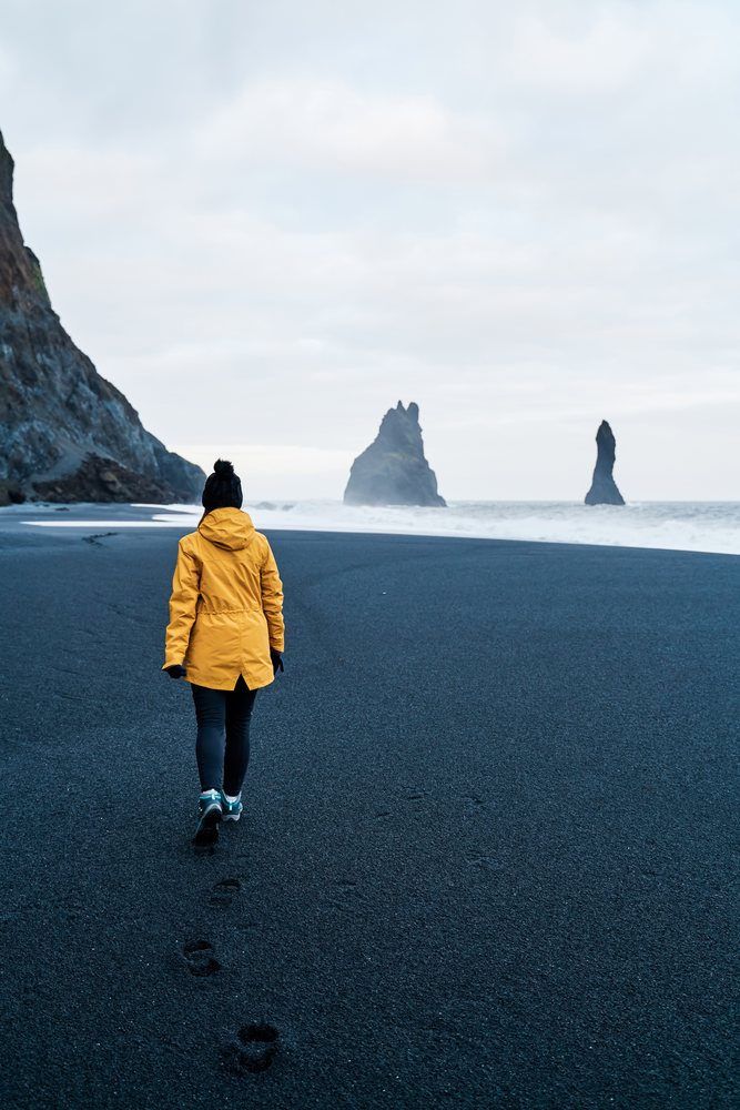 A white person walks on a black sand beach by sea stacks in a beach