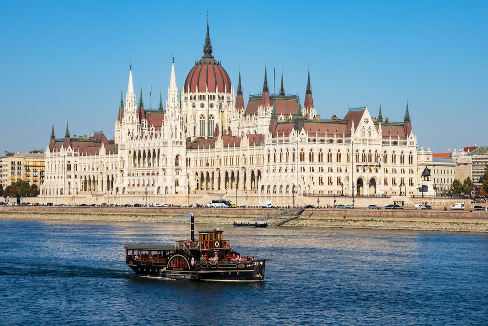 Steam powered paddle wheel boat cruises on the Danube River with the Hungarian Houses of Parliament in Budapest visible along the riverbank under a clear sky, Cityscapes, Hungary.