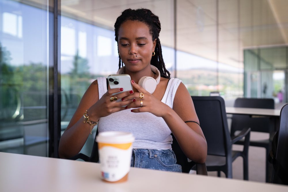 A woman wearing headphones and casual clothing uses her smartphone while sitting at a table with a coffee cup in a modern indoor space, remote work, travel, Santiago De Compostela, Spain.