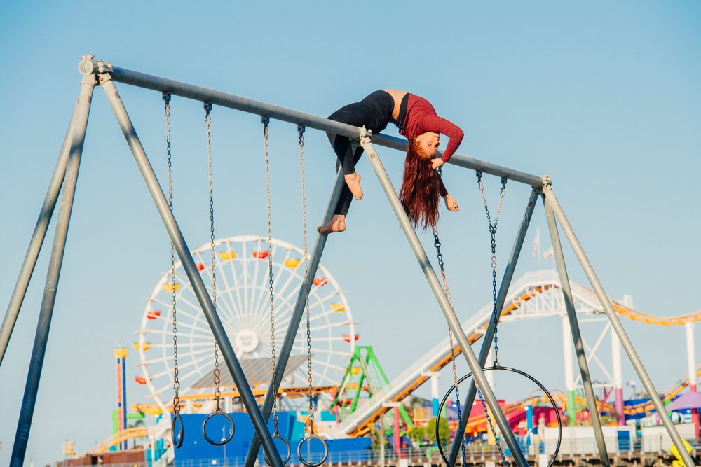 Woman with red hair hangs upside down on metal bars of a swing set at an outdoor amusement park in California under a clear blue sky, USA.