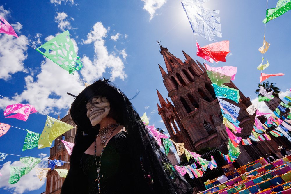 Woman wearing a Calavera mask and black clothing at a Day of the Dead festival in front of a decorated church, San Miguel de Allende, Mexico.