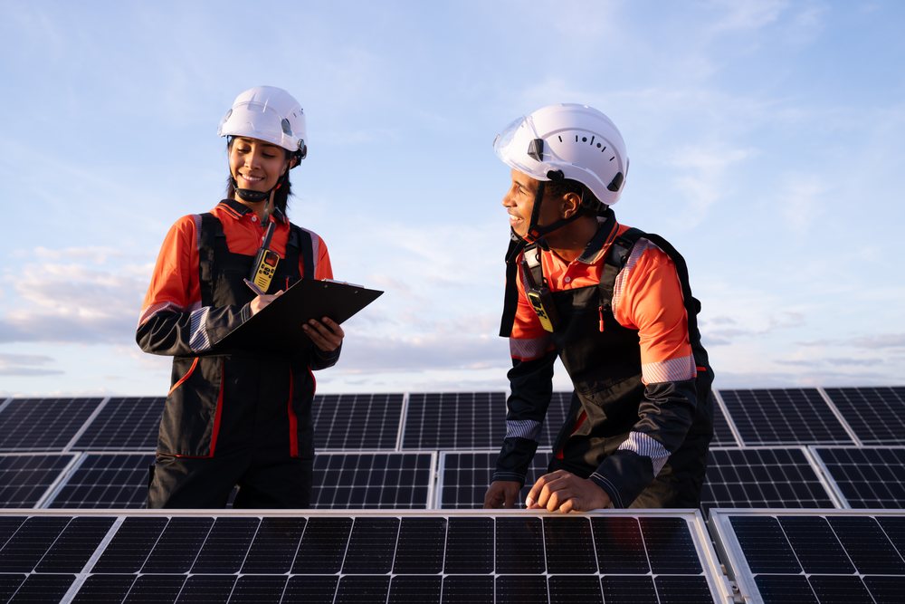 Two engineers wearing protective helmets and communication devices inspect solar panels outdoors at sunset, Madrid, Spain.