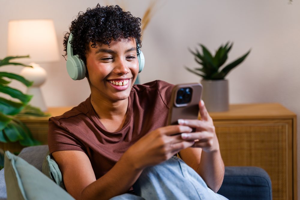 Woman with curly hair wearing wireless headphones uses a smartphone while sitting on a couch in a living room with decorative plants and a lamp, Spain.