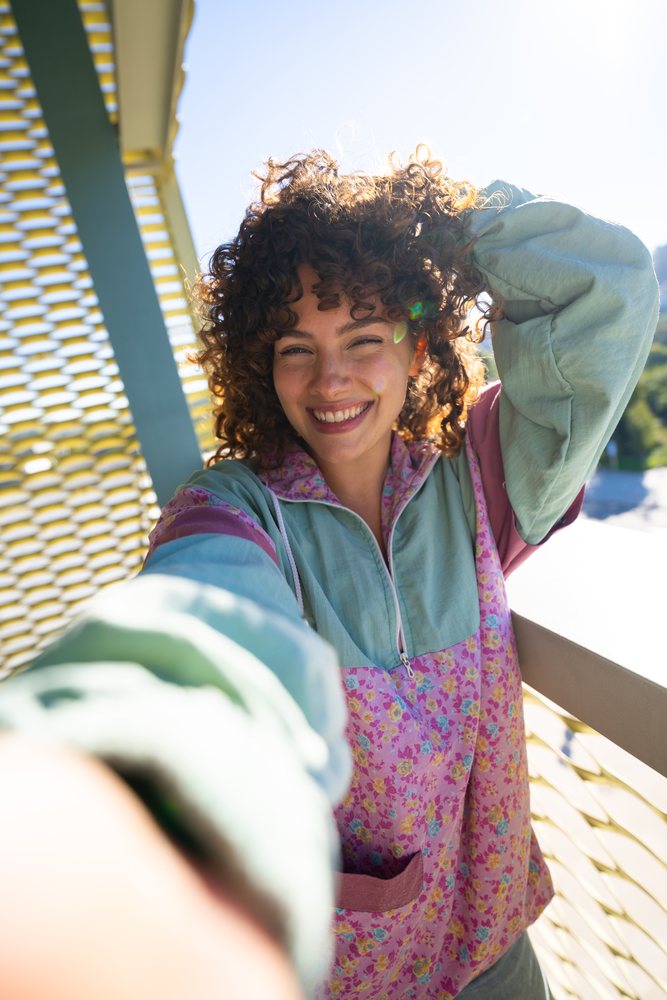 Woman with curly hair wearing a colorful floral jacket smiles while taking a selfie outdoors in sunlight by a mesh fence, Spain.