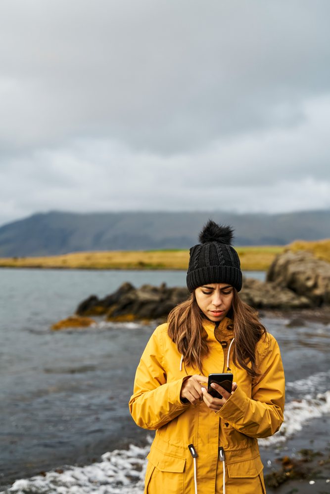 A white woman uses her smartphone by the rocky shore