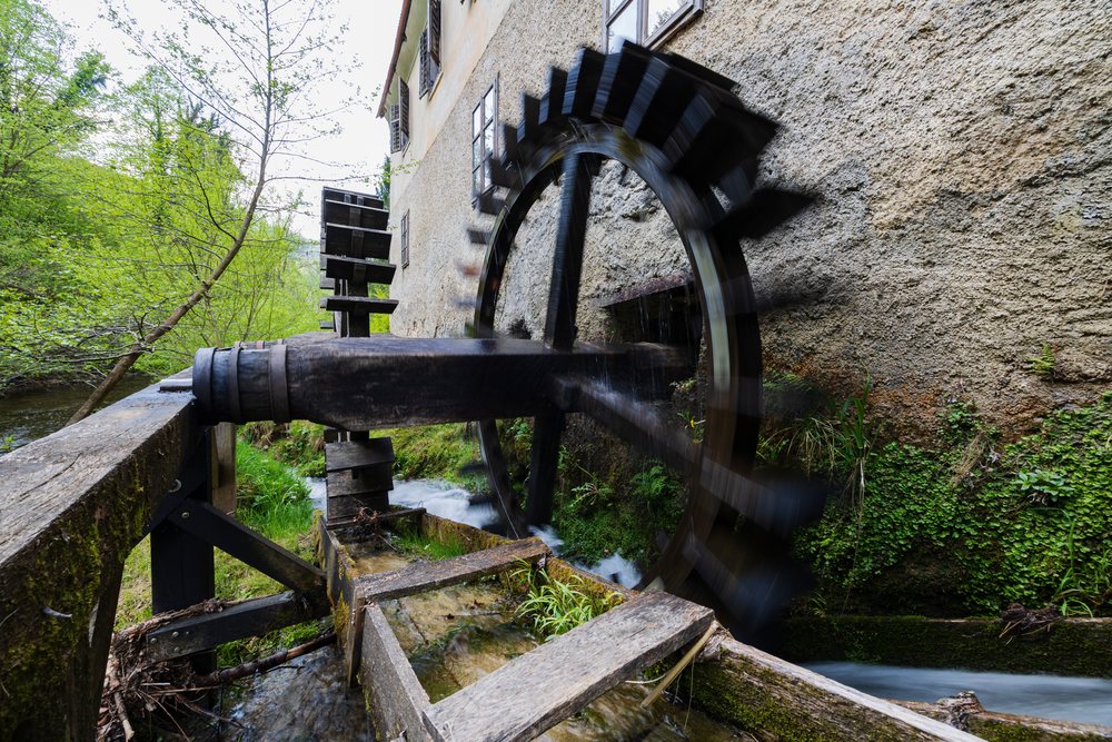 Water wheel turning beside the historic Kukovičičev Mlin mill, with flowing water and green foliage nearby, landscape, cityscape, Podsreda, Slovenia.