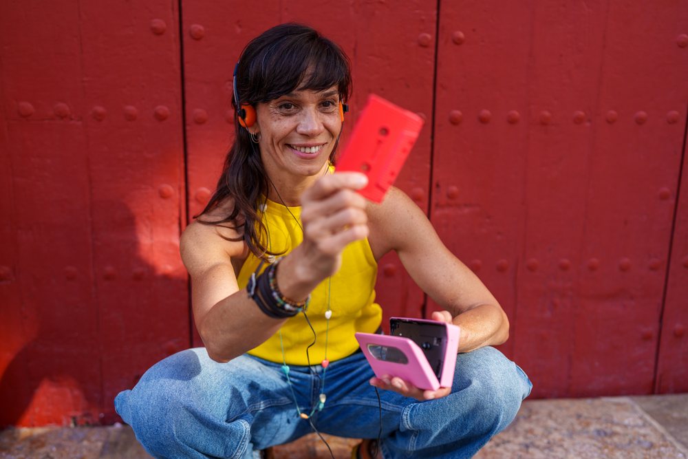 Woman sitting outdoors holding a pink cassette player and red cassette tape near a red door, retro, vintage, Málaga, Spain.