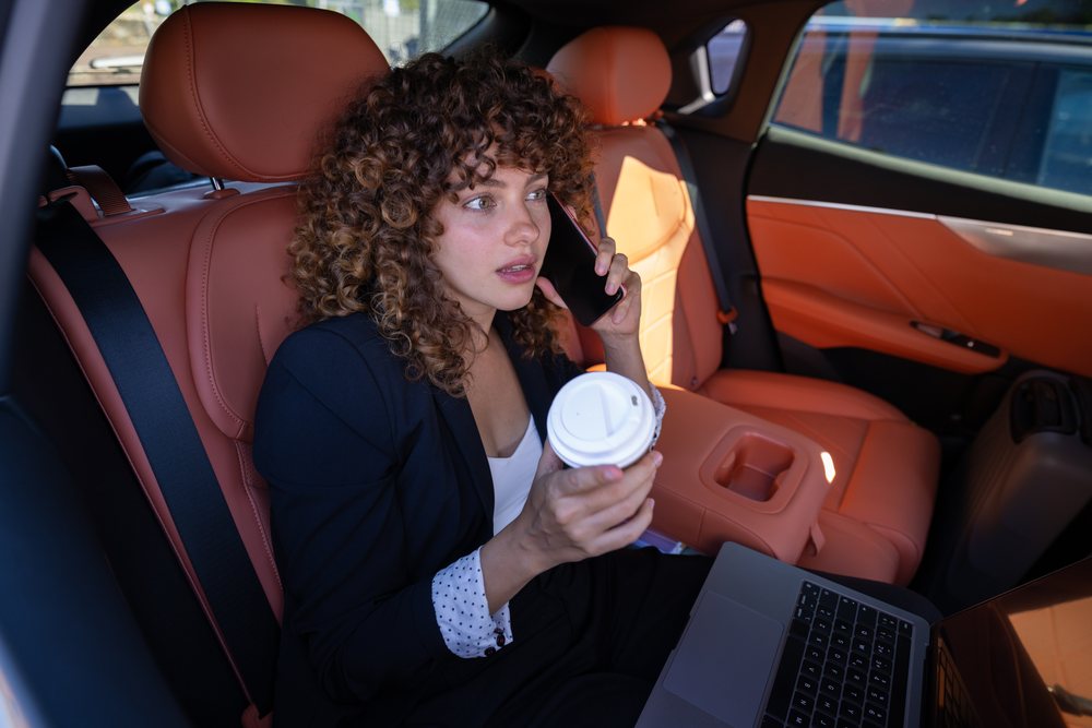 Woman with curly hair talks on a mobile phone and holds a coffee cup while sitting inside an orange leather-seated car with a laptop on her lap, remote work, businesswoman, Spain.