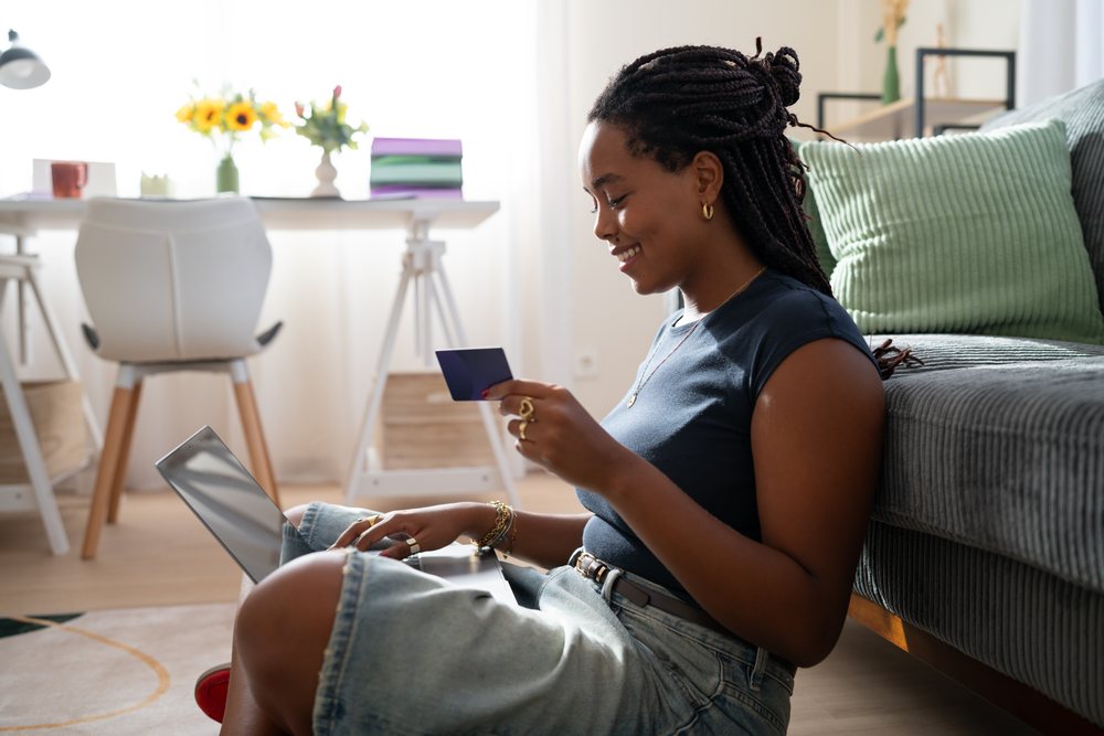 A woman smiles while sitting on the floor and using a laptop with a credit card in a well-lit home office setting, remote work, student, Spain.