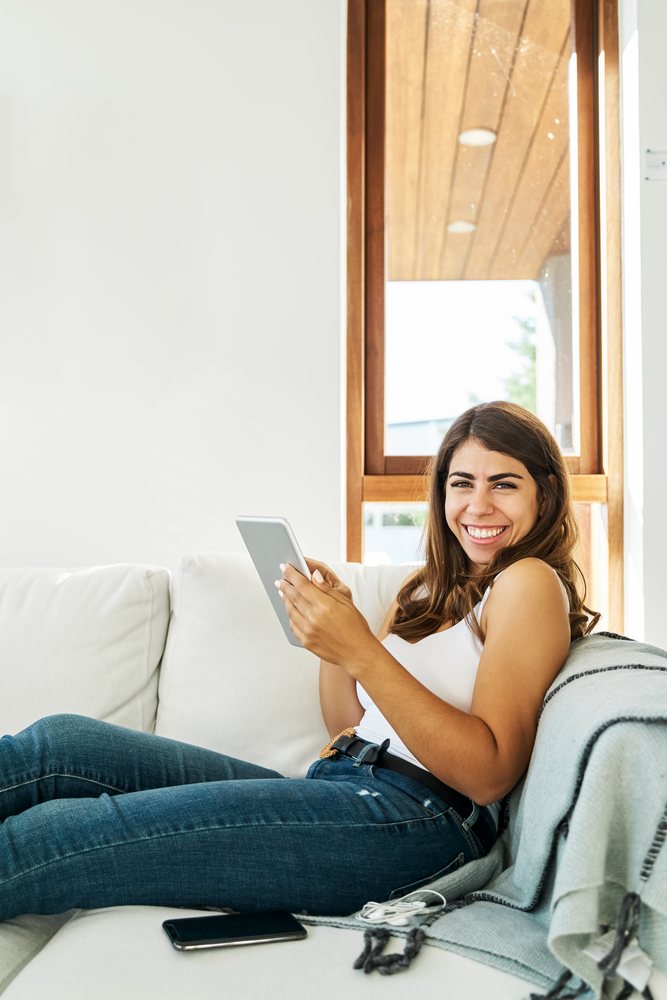 A smiling white woman relaxes on a sofa using a tablet
