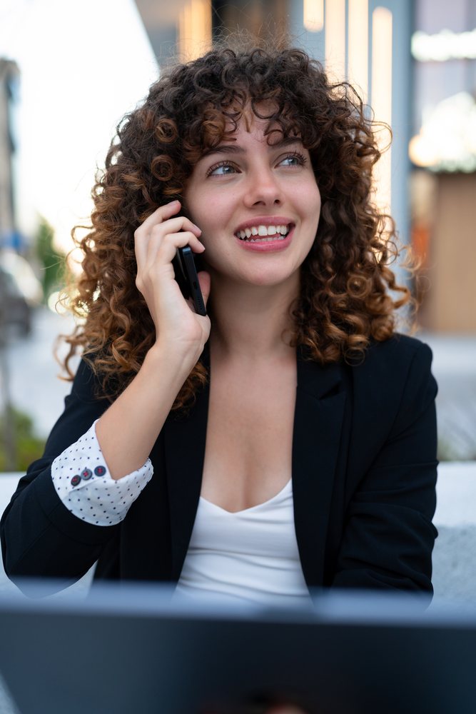 Young woman with curly hair seated outdoors, smiling while talking on phone, Madrid.